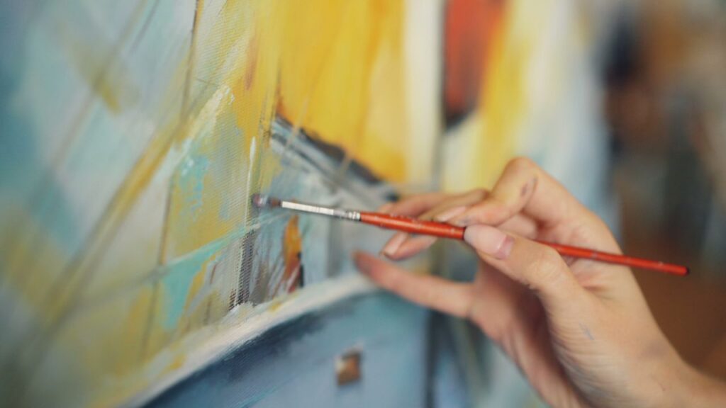 Close-up of an artist's hand using a brush to paint on a colorful canvas.